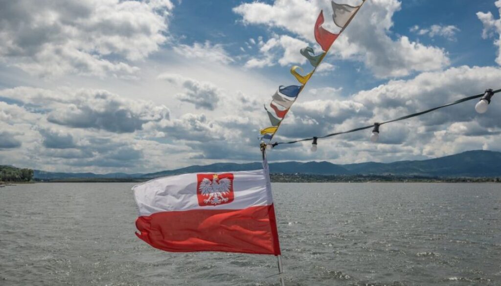 Atrakcje turystyczne na jeziorach w Beskidach. Na zdjęciu widoczna jest polska flaga (rejst statkiem turystycznym po Jeziorze Żywieckim) na tle tafli jeziora w górach. Z samego tyłu widoczne są Beskidy, dokładnie SKrzyczne w Beskidzie Śląskim.