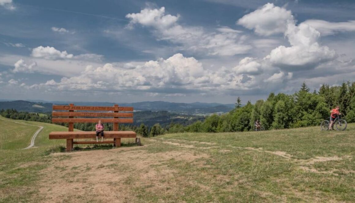 Największa ławka zakochanych na Słowacji znajduje się na Trojaku, czyli na Kikuli nad Zwardoniem. Na zdjęciu widoczna kobieta na wielkiej ławce na Trojaku. Tło dla zdjęcia stanowią góry w Beskidach. Zdjęcie wykonane latem w góach, z zieloną trawą i pięknymi chmurami na niebie.